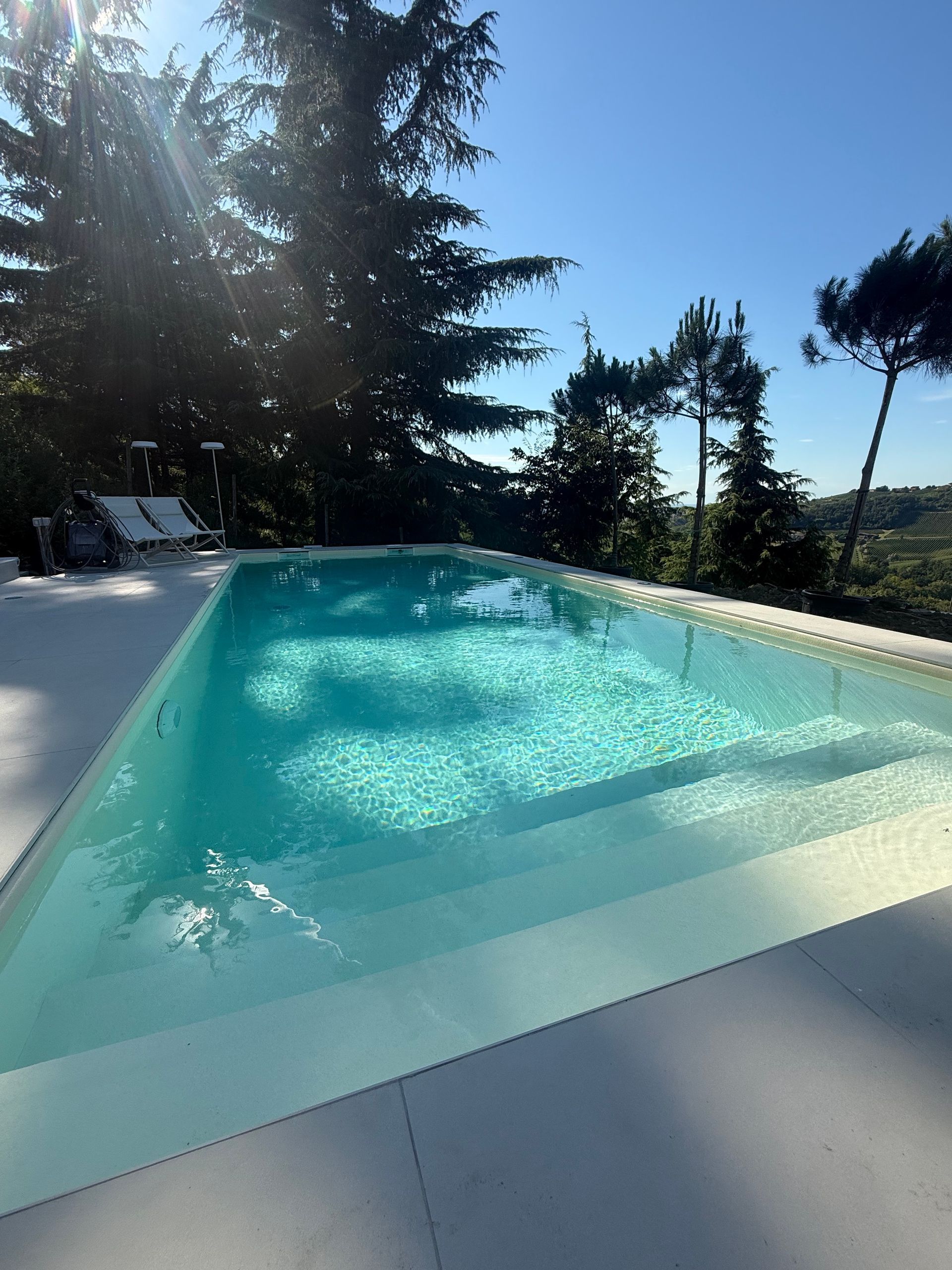 Swimming pool with clear turquoise water, surrounded by white concrete. Sunlight streams through trees.