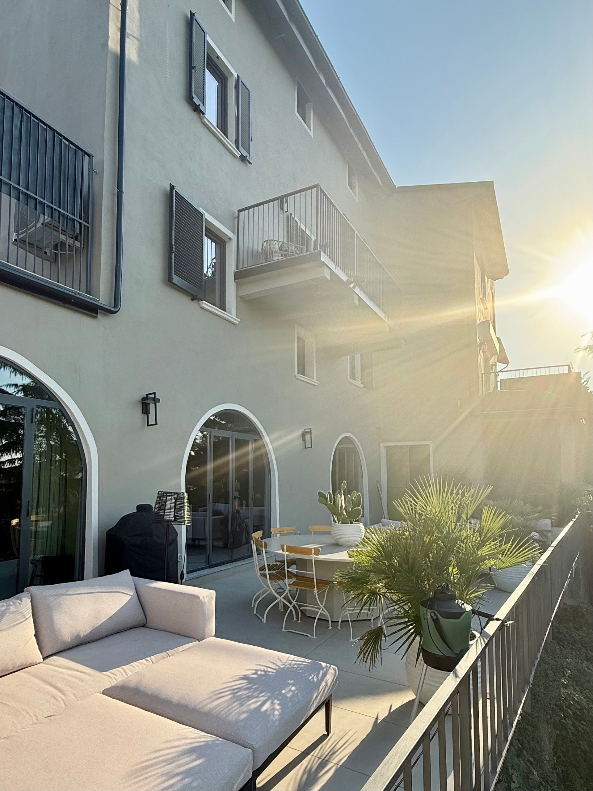 Outdoor patio with a beige sectional, table, and arched doorways. Bright sunlight.