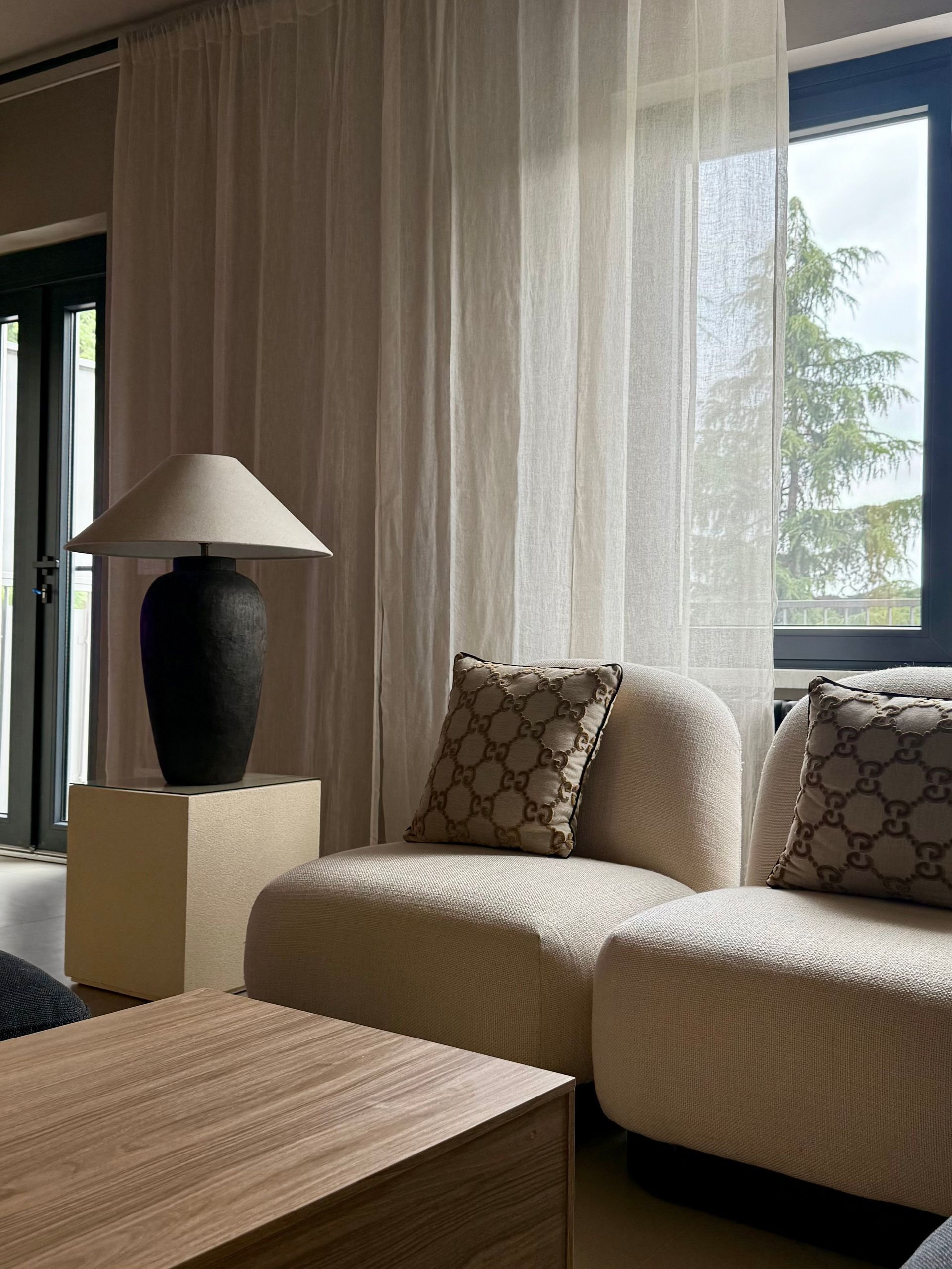 Cozy living room with cream-colored sofa and Gucci pillows. A black lamp sits on a beige cube. Sheer curtains filter light.