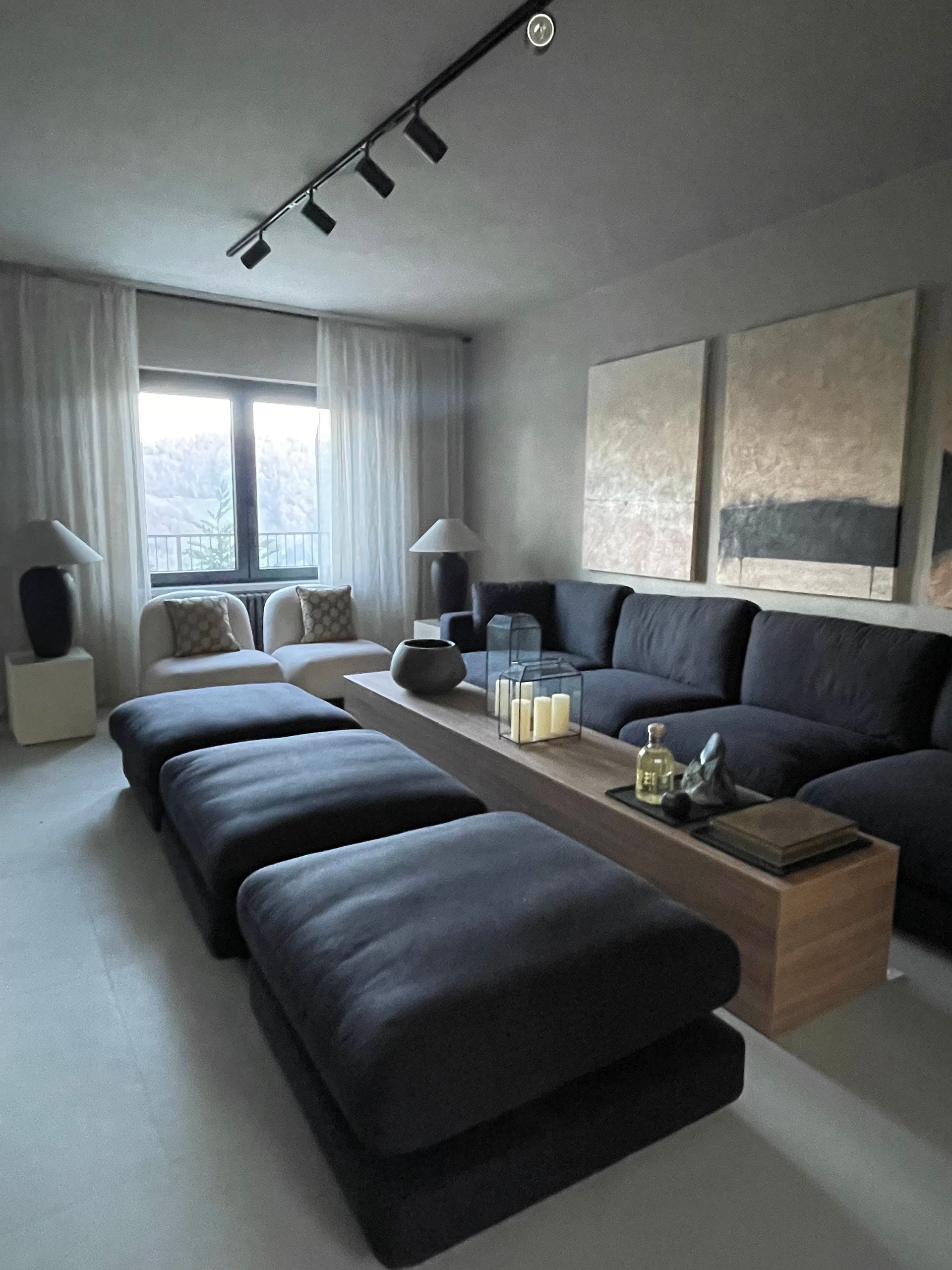 Modern living room with dark gray sectional, ottomans, light-colored curtains, and artwork.