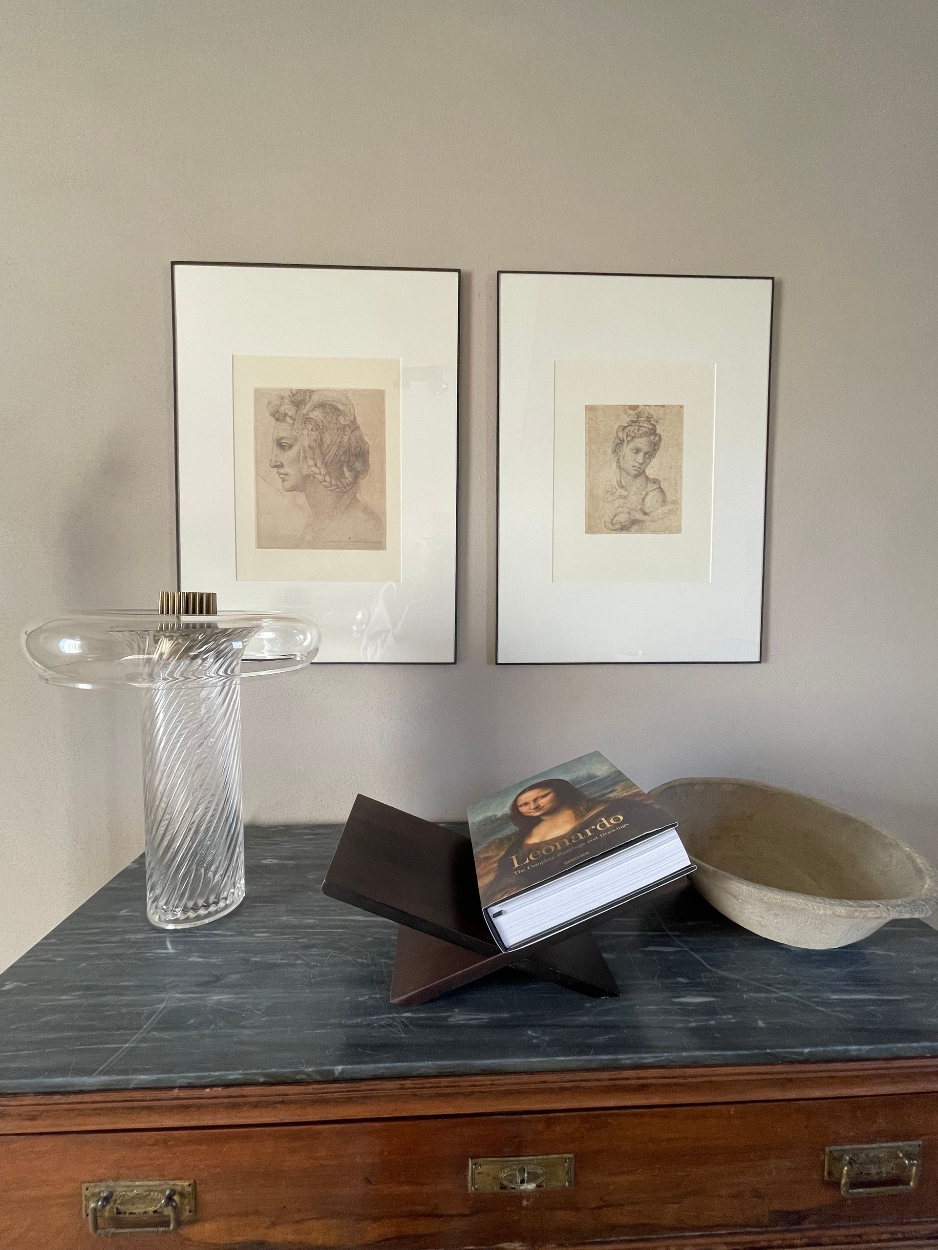 Two framed drawings and objects on a dresser. A glass vase, a book, and a woven bowl.