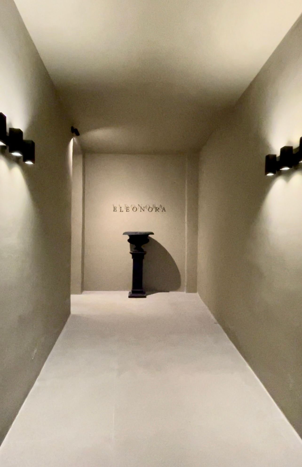 Narrow beige hallway with a black pedestal, sconces on each side, and  ELEONORA on the back wall.