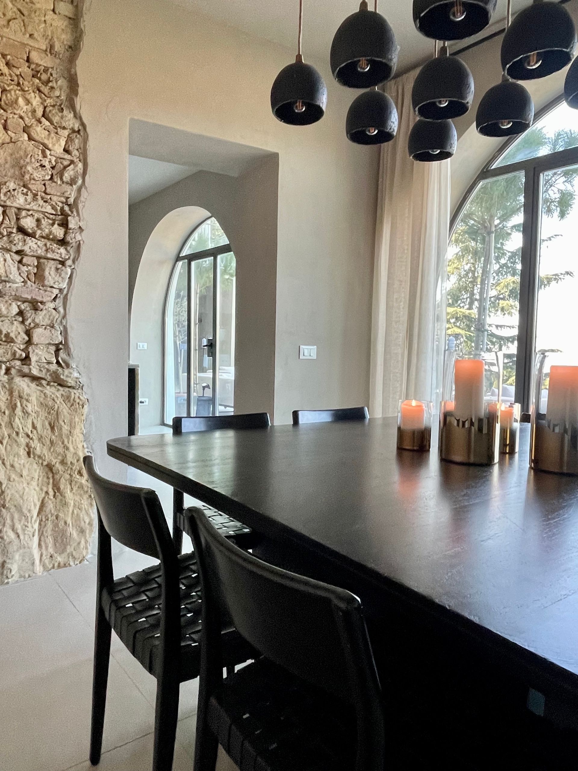 Dining room with long dark table, black chairs, hanging lamps, candles, and archway to another room.