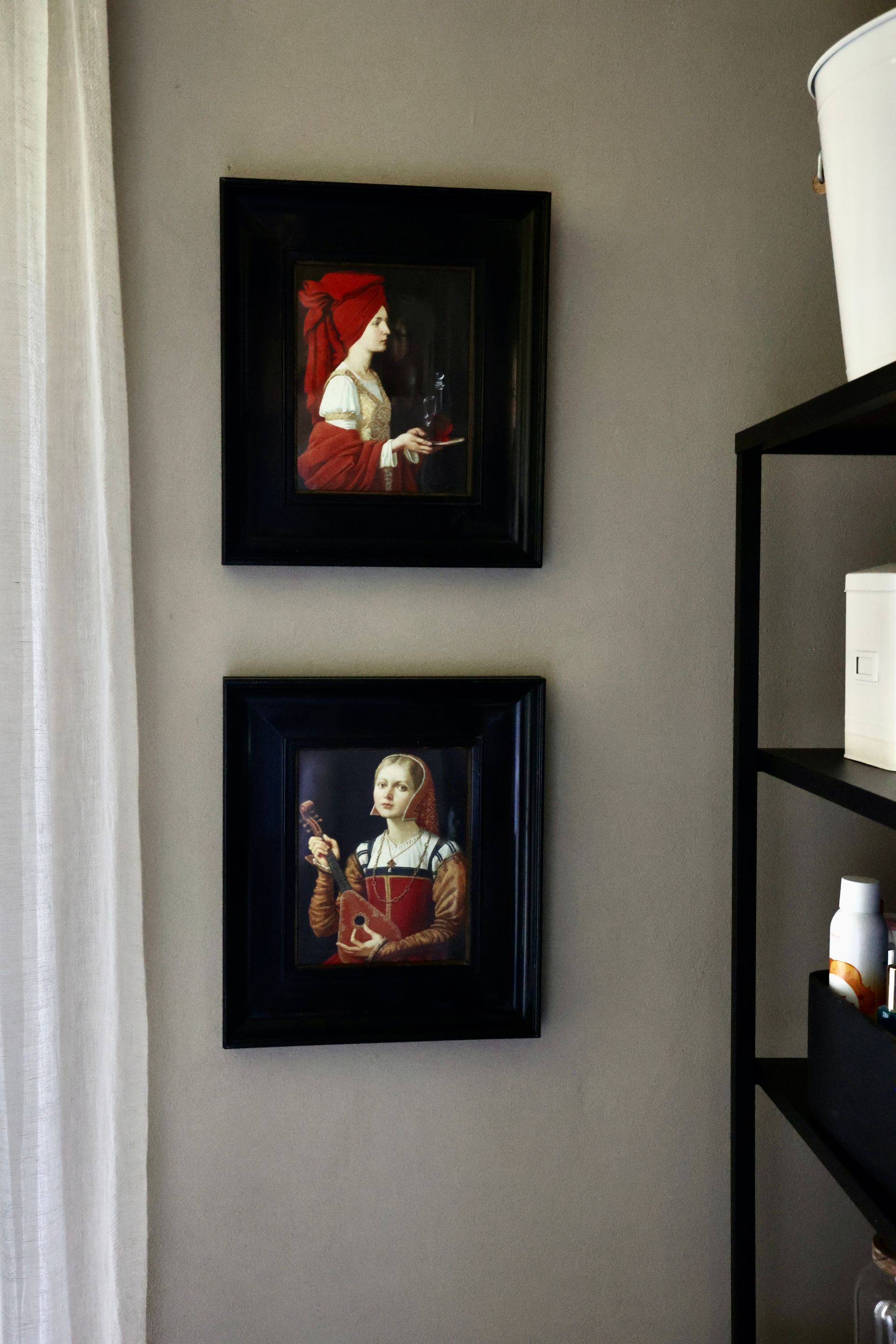 Two framed portraits hang on a gray wall, with a sheer curtain on the left and shelving on the right.