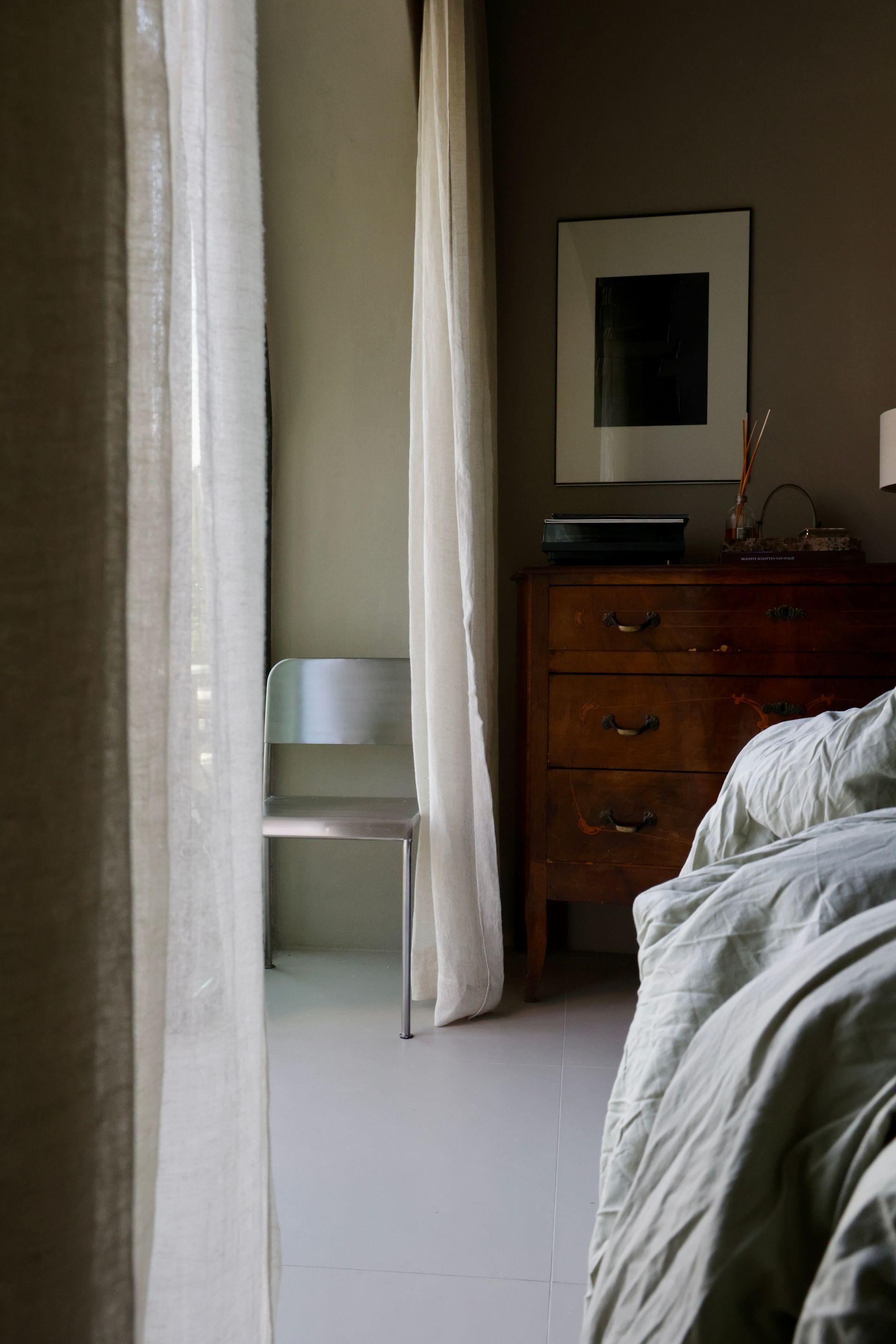 Bedroom interior with sheer curtains, wooden dresser, bed, and chair.