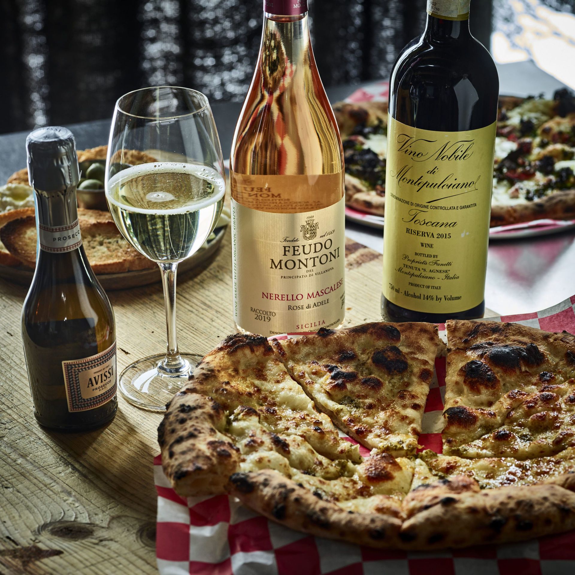 Pizzas, wine bottles, and a glass of wine on a table with a checkered napkin.