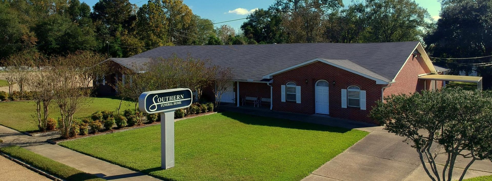 A brick building with a dark roof and a sign in front, surrounded by a green lawn and trees.
