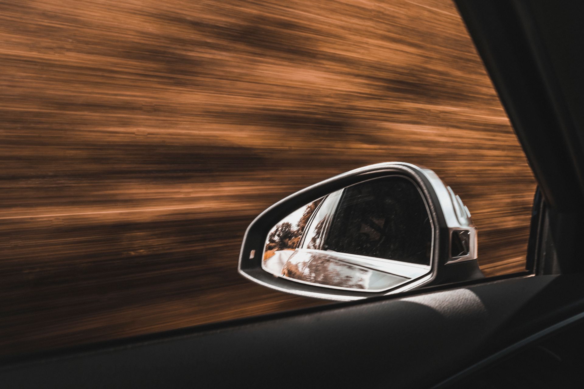 Side-view mirror of a moving car, reflecting scenery blurred by speed, earthy brown tones.