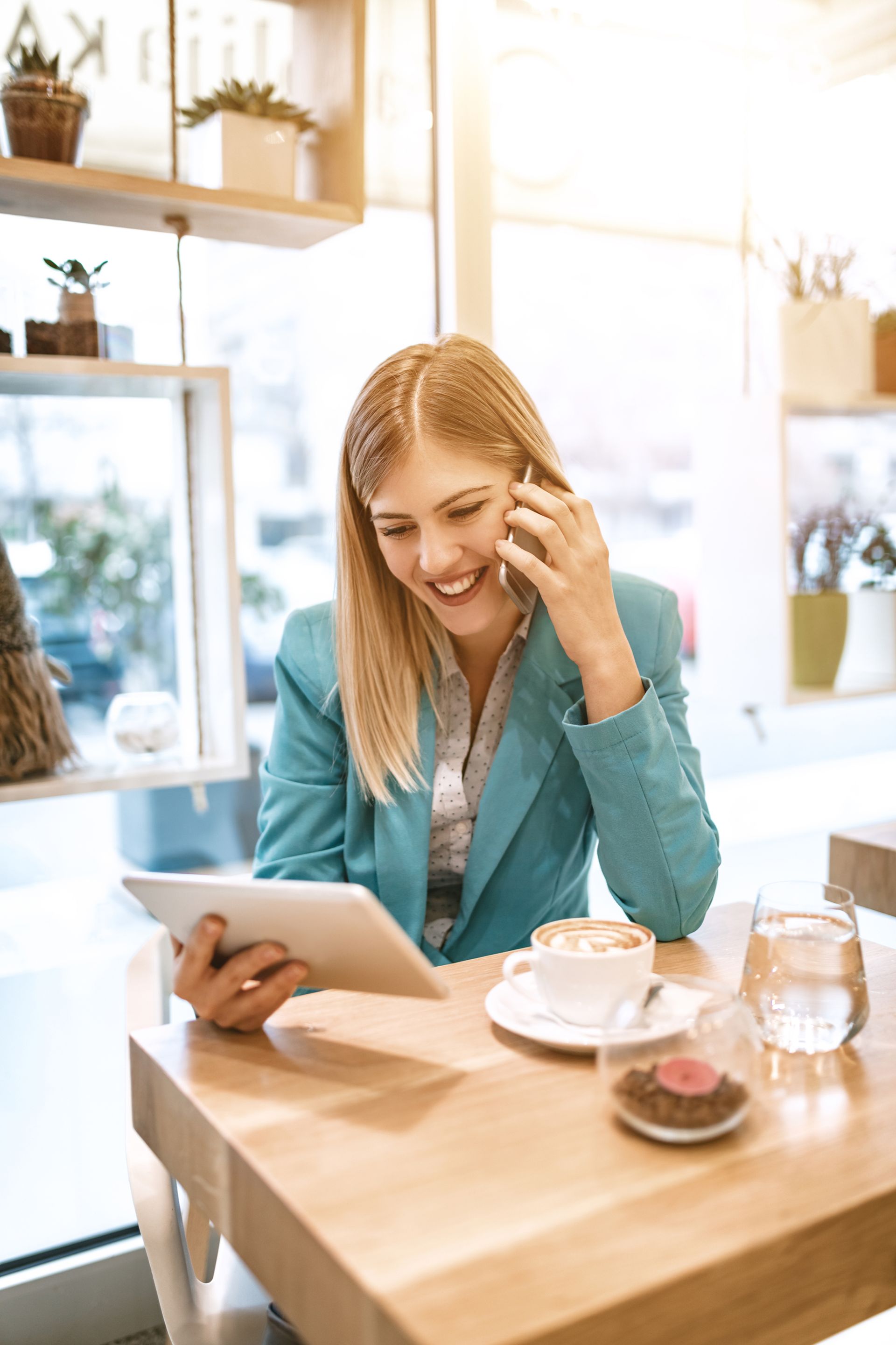 Woman in blue blazer smiles while on phone and using a tablet in a cafe, with coffee and water on the table.