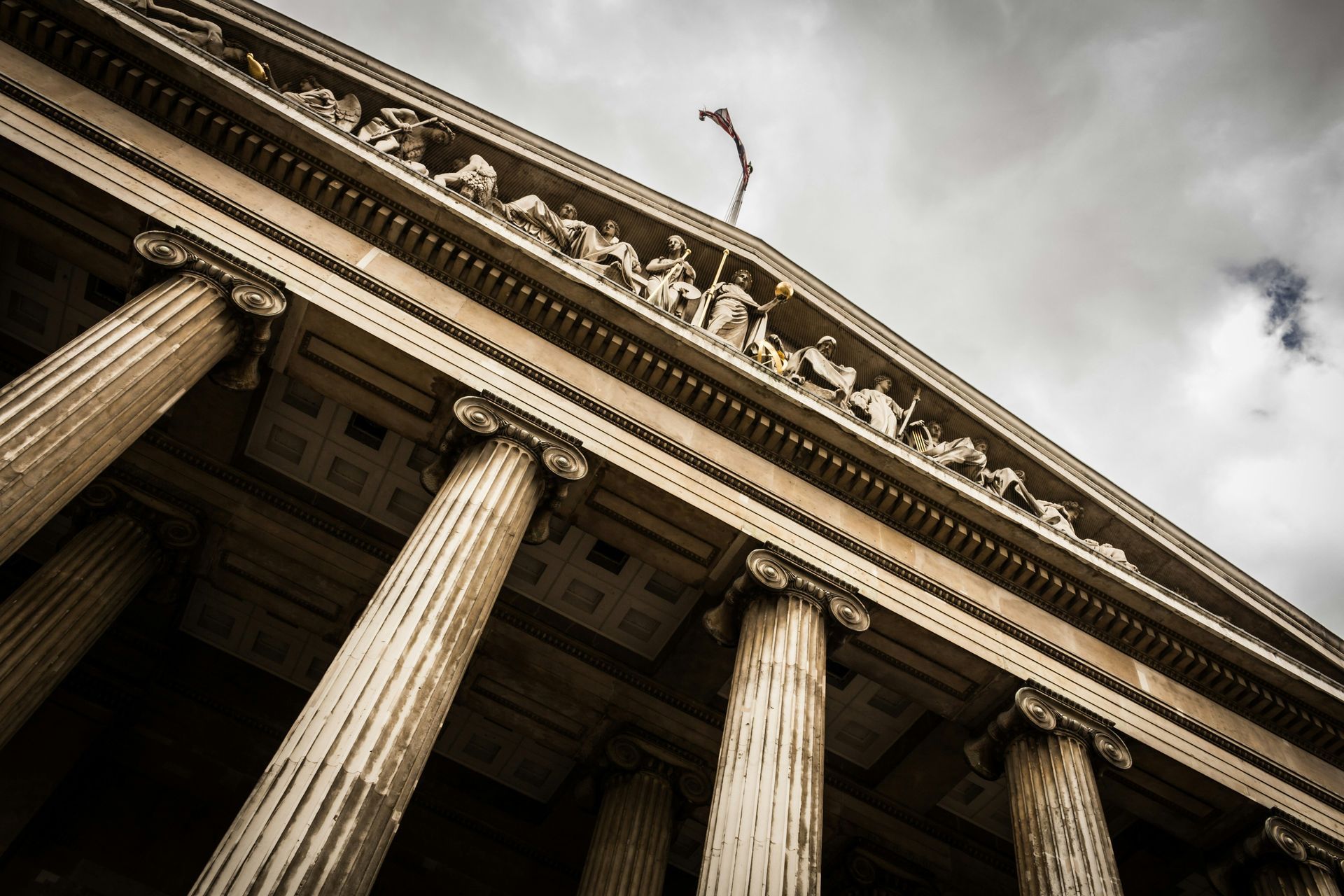 Low-angle view of a classical building with columns, sculptures, and a cloudy sky.