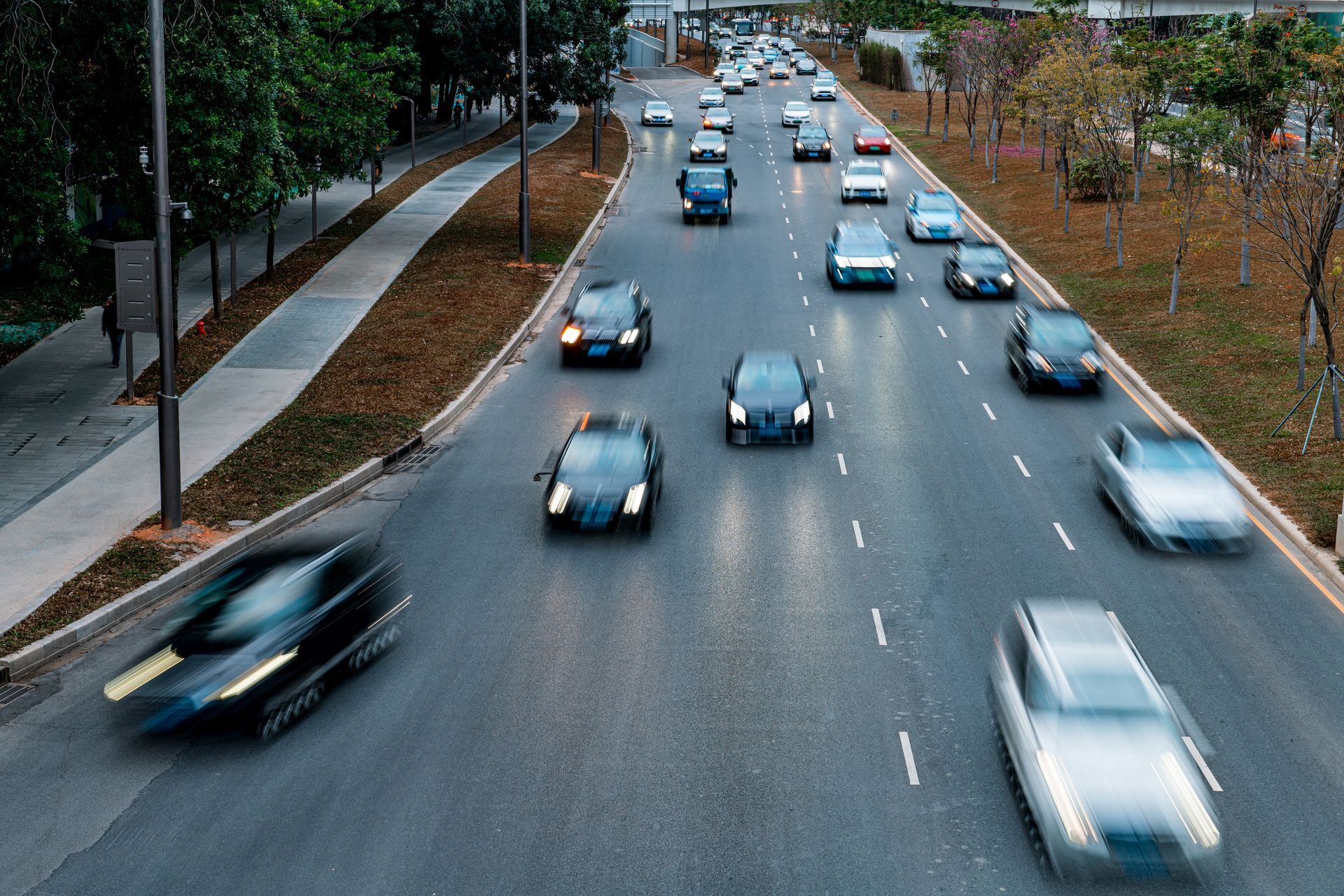Cars traveling on a multi-lane highway, asphalt road with white lane markings, trees and sidewalk alongside.