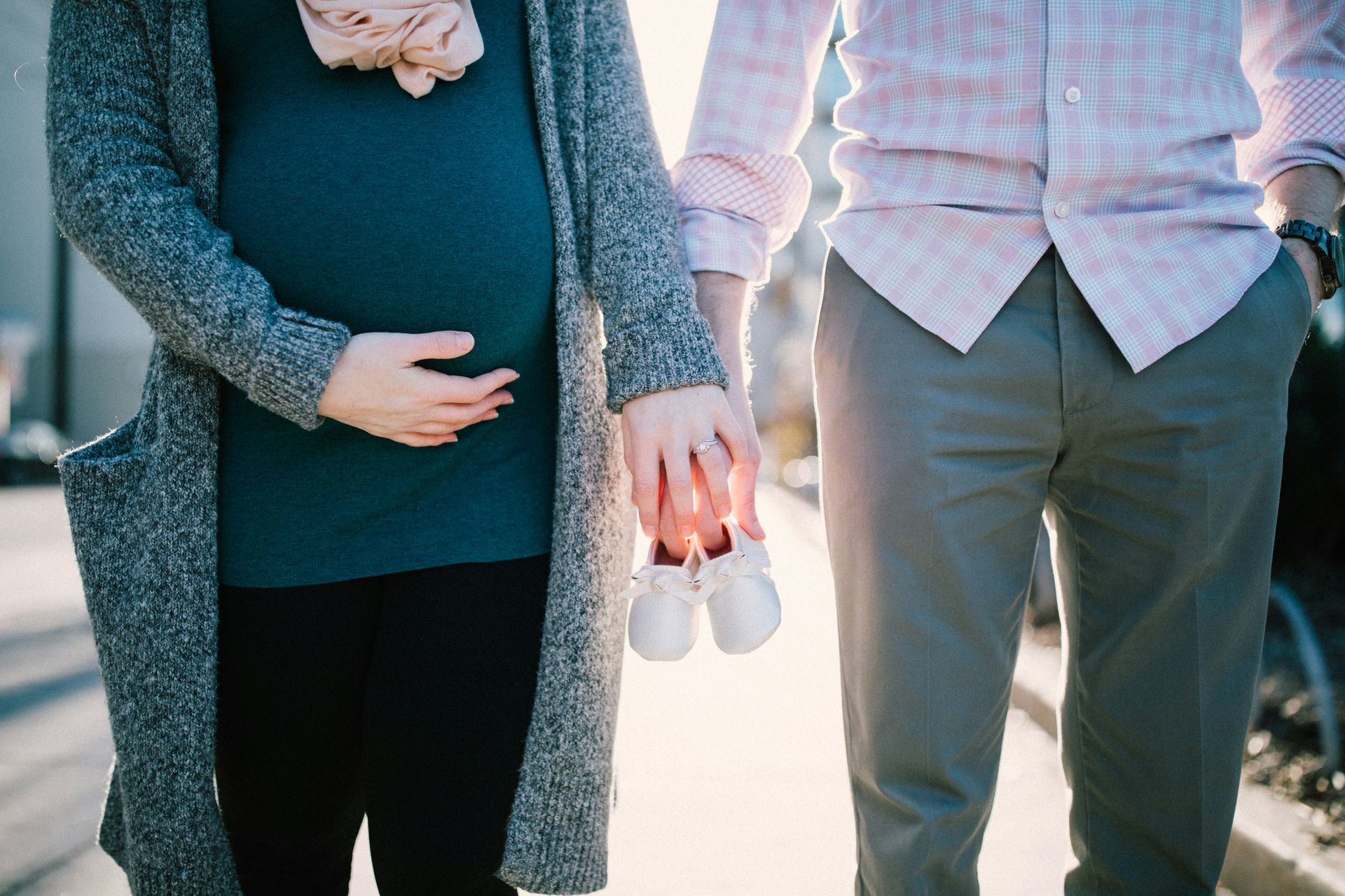Pregnant woman and partner hold hands and baby shoes, outdoors.