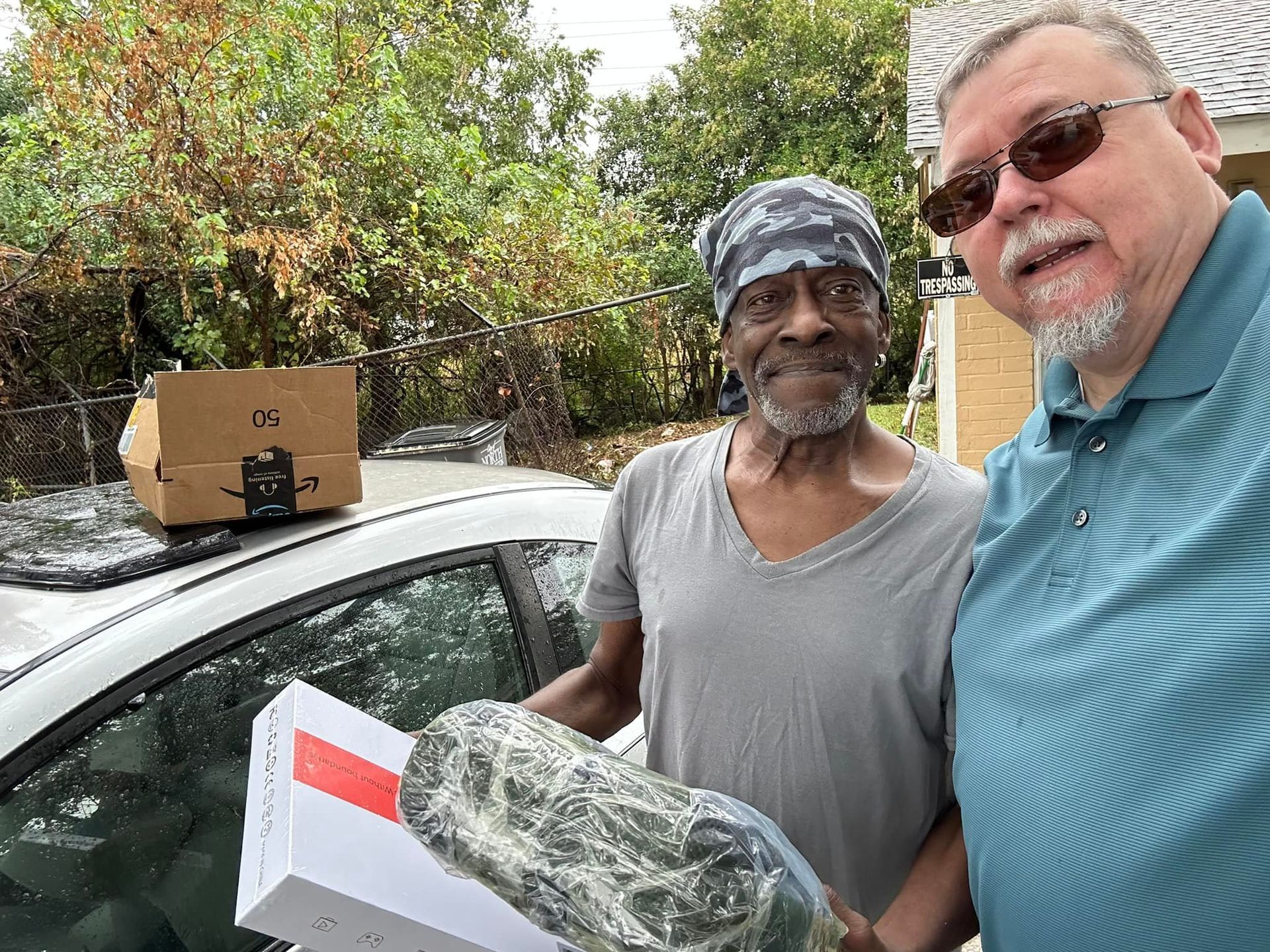 Two men are standing in front of a car holding boxes.