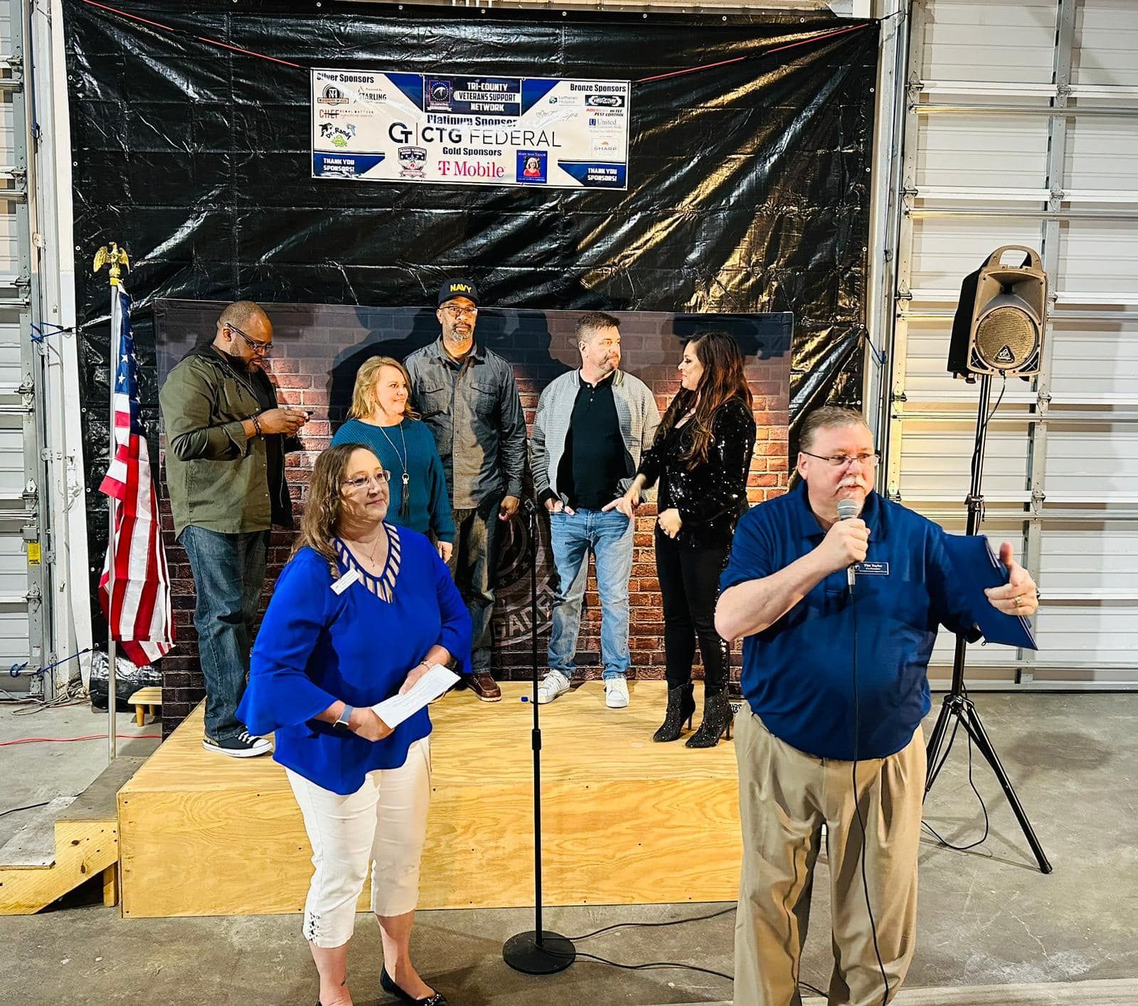 A group of people are standing on a stage with a man speaking into a microphone.