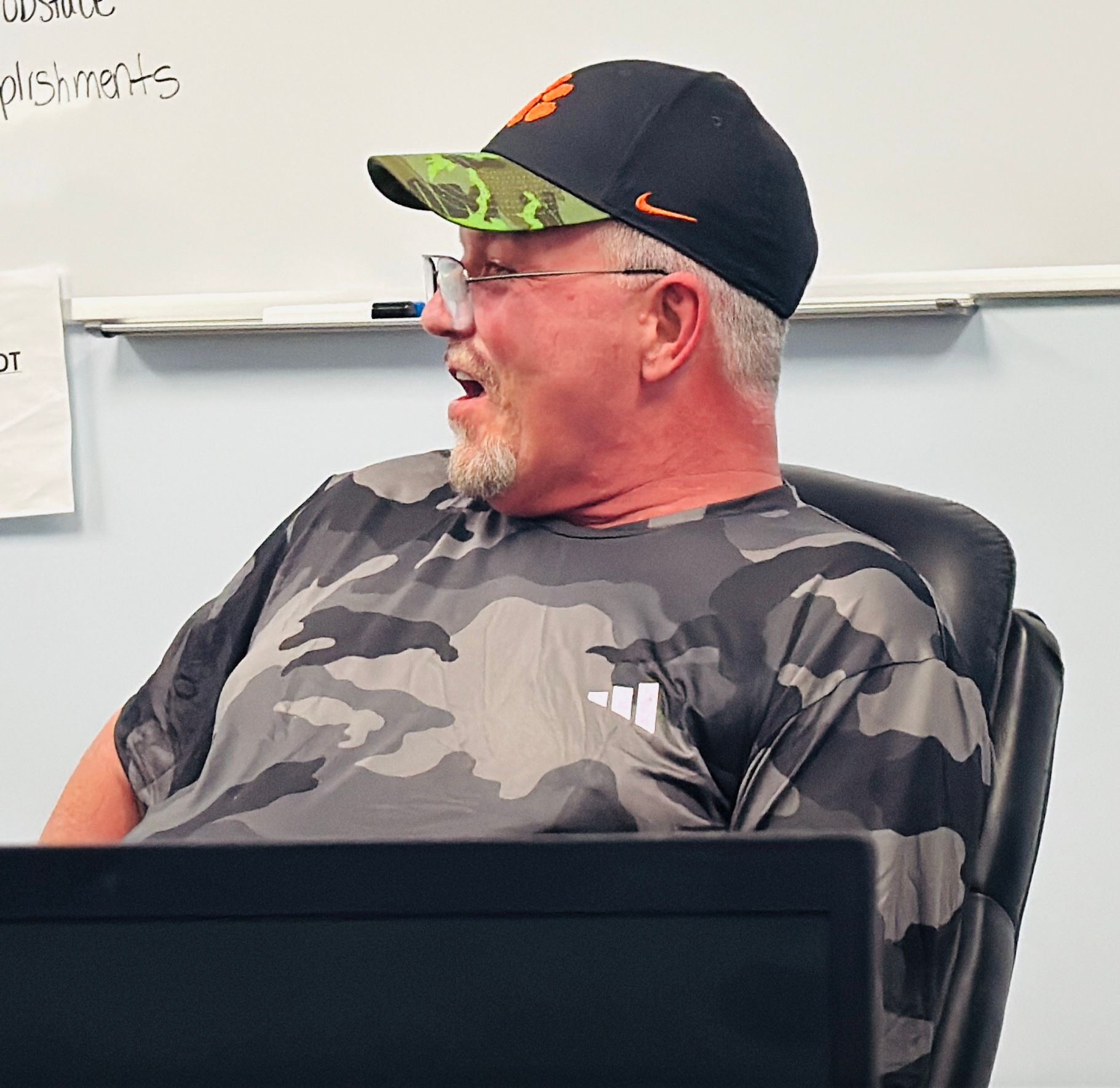 A man wearing a hat and glasses is sitting in front of a white board with the word shirts written on it