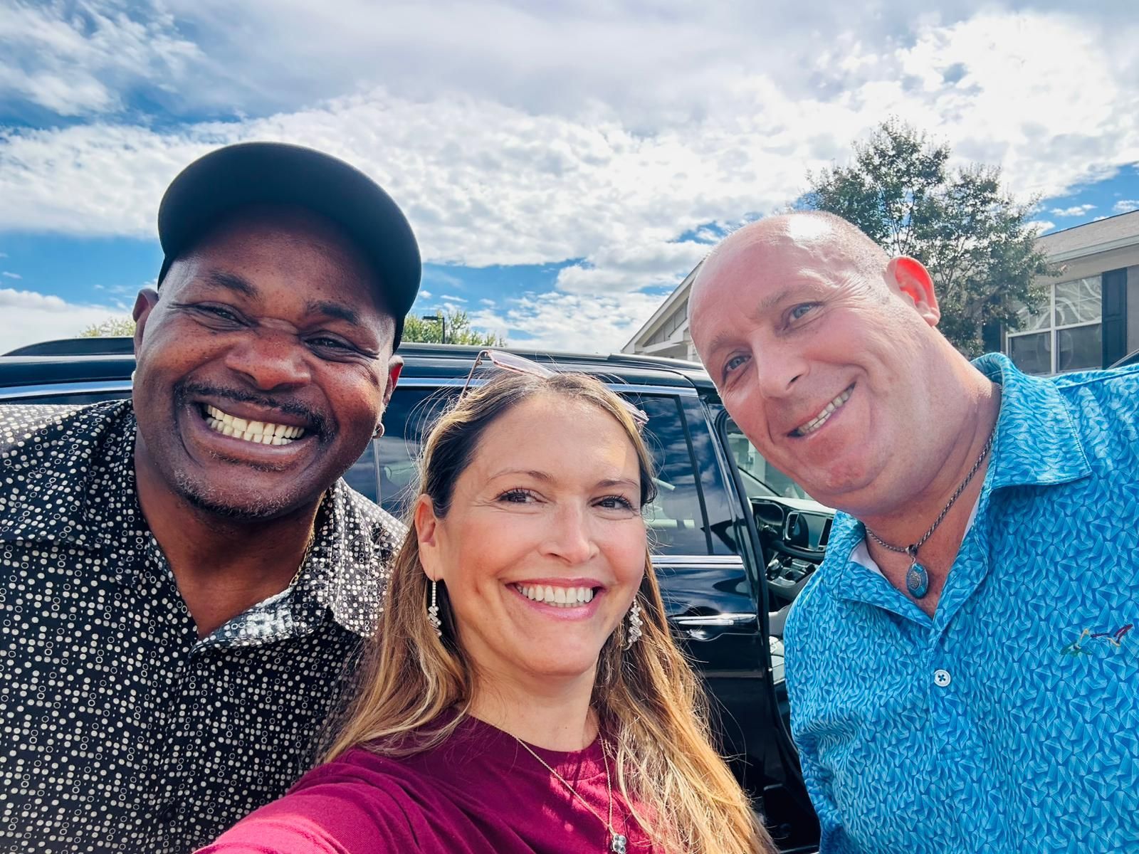Two men and a woman are posing for a picture in front of a car.