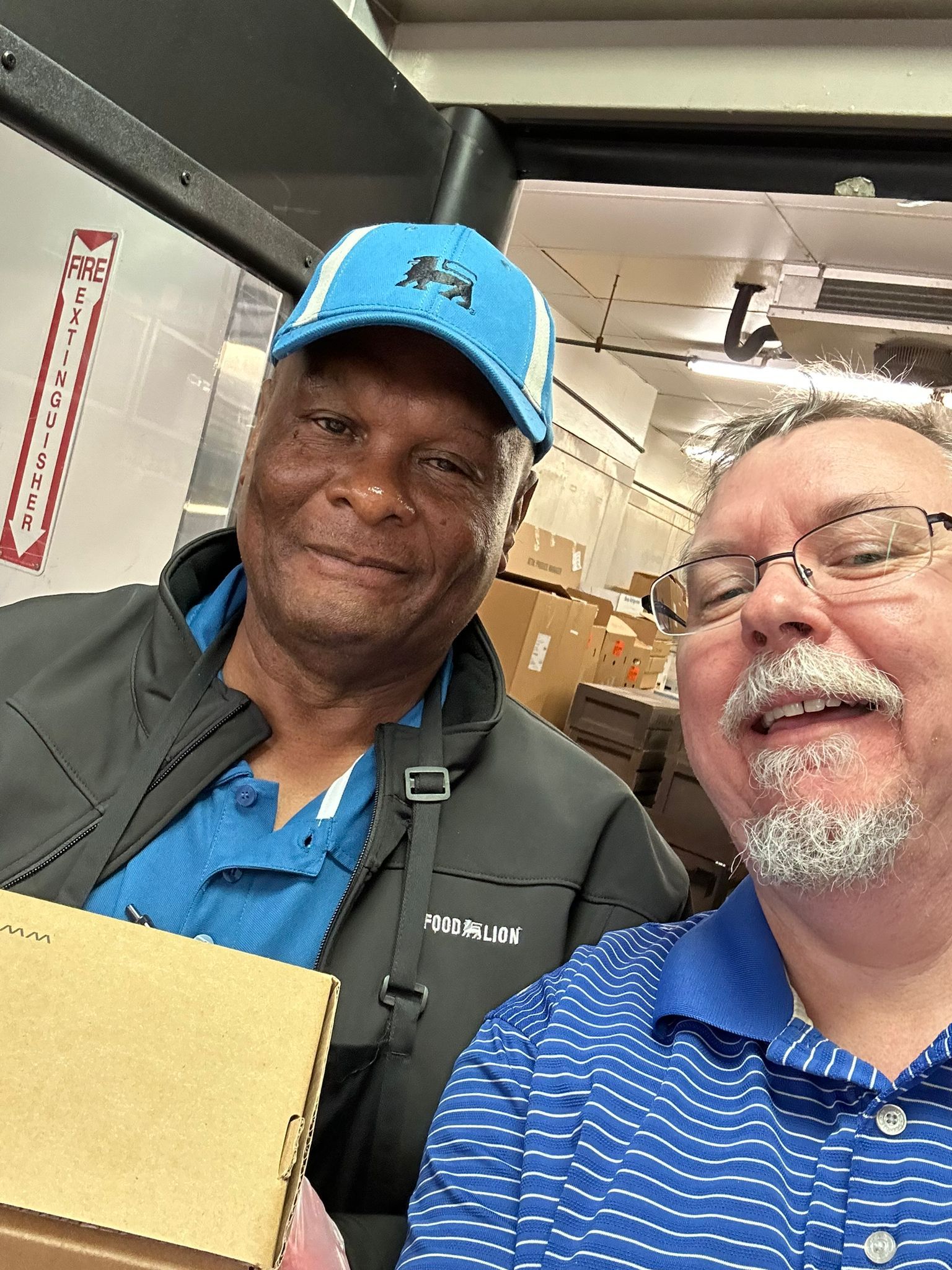 Two men are posing for a picture in a warehouse while one of them is holding a box.