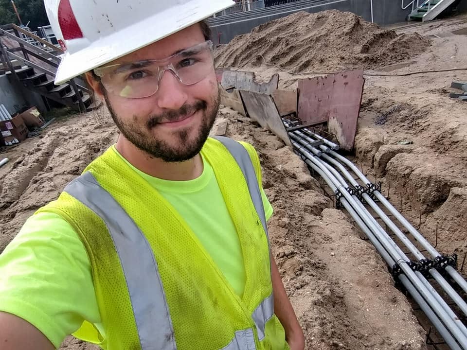 A man wearing a hard hat and safety vest is standing in the dirt.