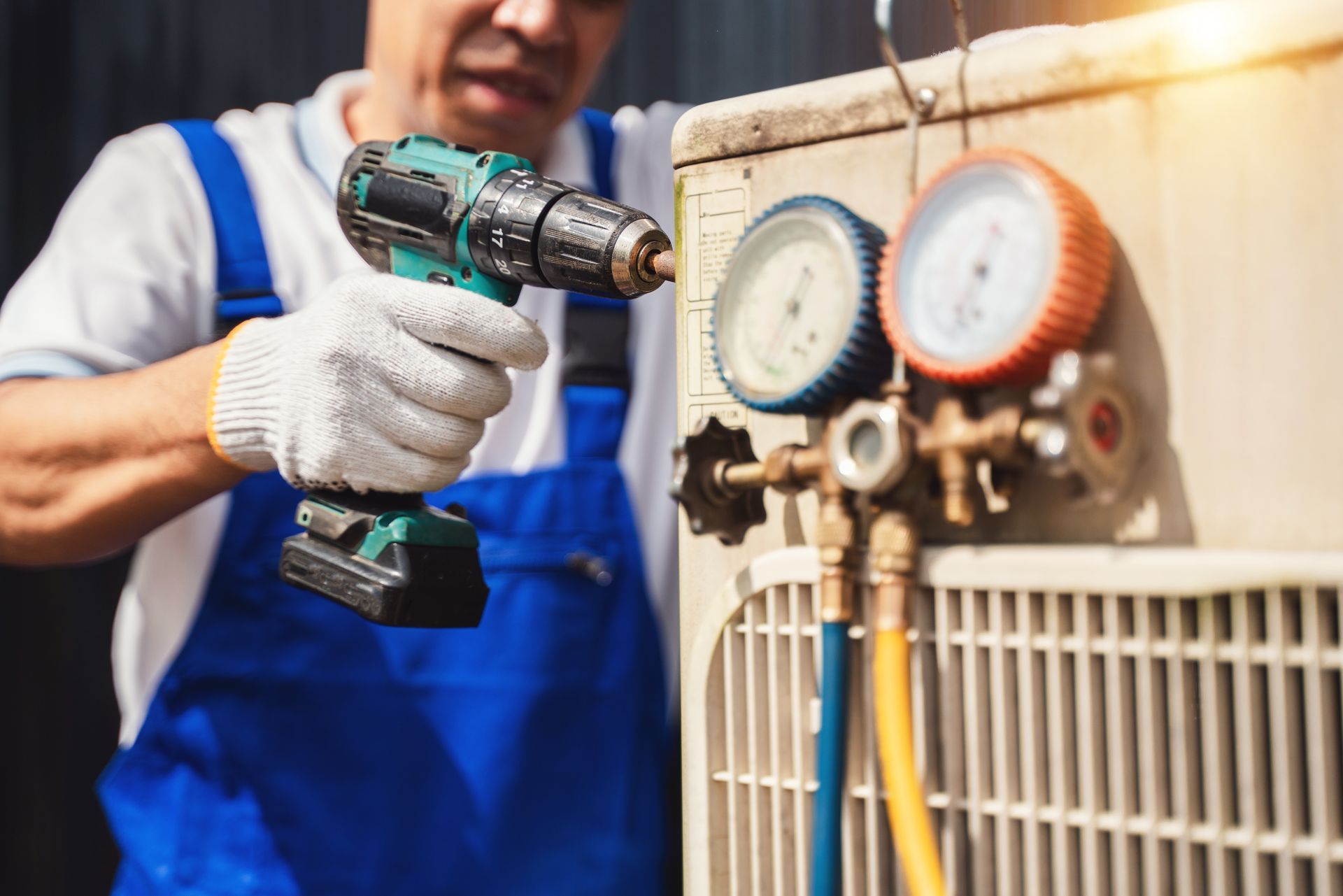Technician in blue overalls using a cordless drill to service an HVAC unit with gauges attached. Technician in blue overalls using a cordless drill to service an HVAC unit with gauges attached.