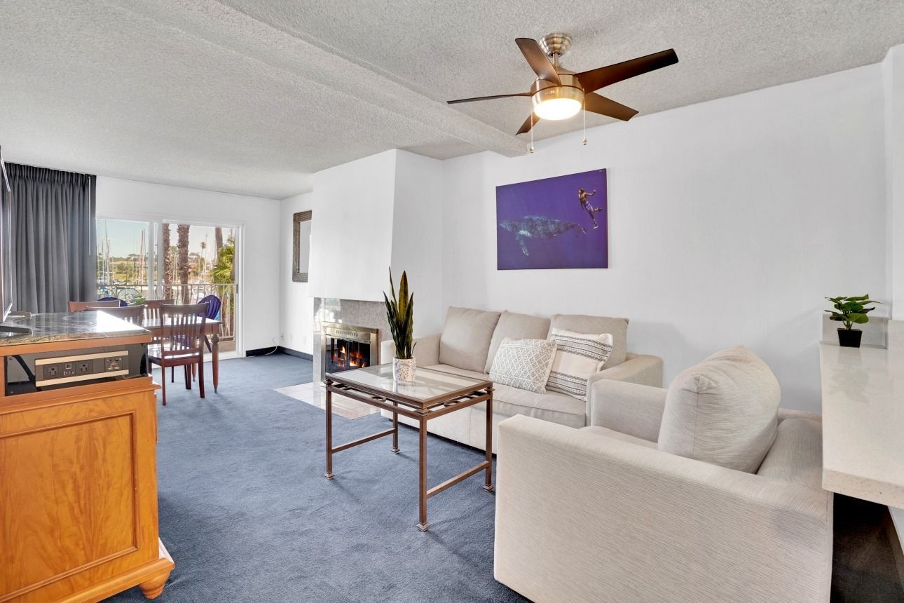 A living room with blue carpet, a beige sofa and armchair, a coffee table, a ceiling fan, and a view to a dining area.