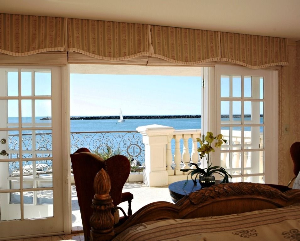 A bedroom window view overlooking a balcony and the ocean with a sailboat in the distance.