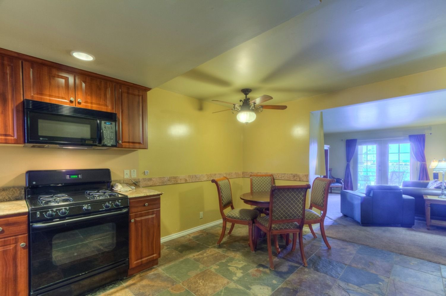 A kitchen with wood cabinets, black appliances, and stone flooring, opening into a living room with a sofa and patio doors.
