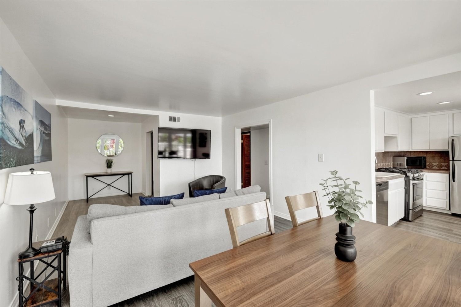 A bright, modern open-plan living and dining area featuring a light gray couch, wooden dining table, and white kitchen.