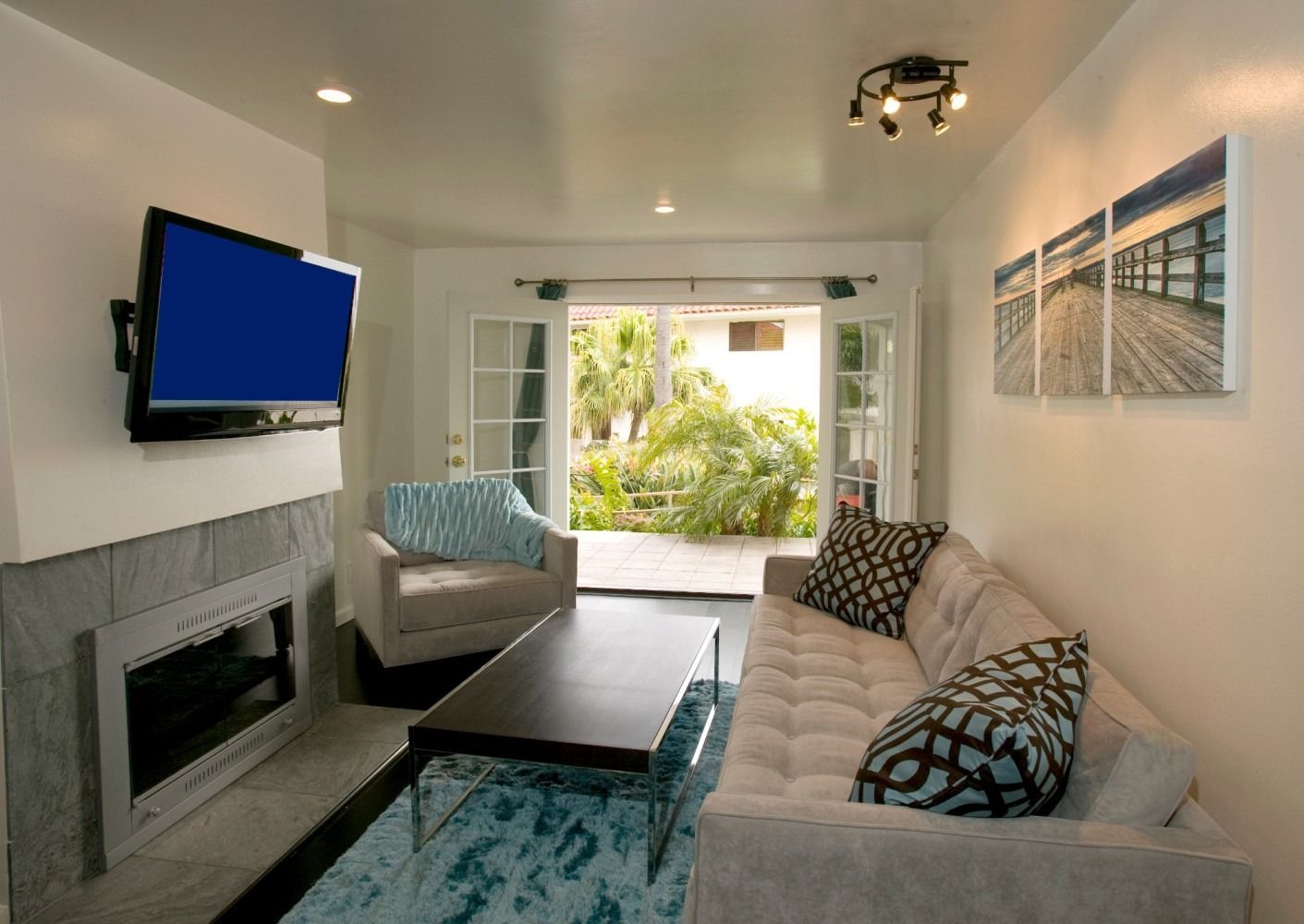 A contemporary living room with a television above a fireplace, a sofa, a coffee table, and open French doors to a patio.
