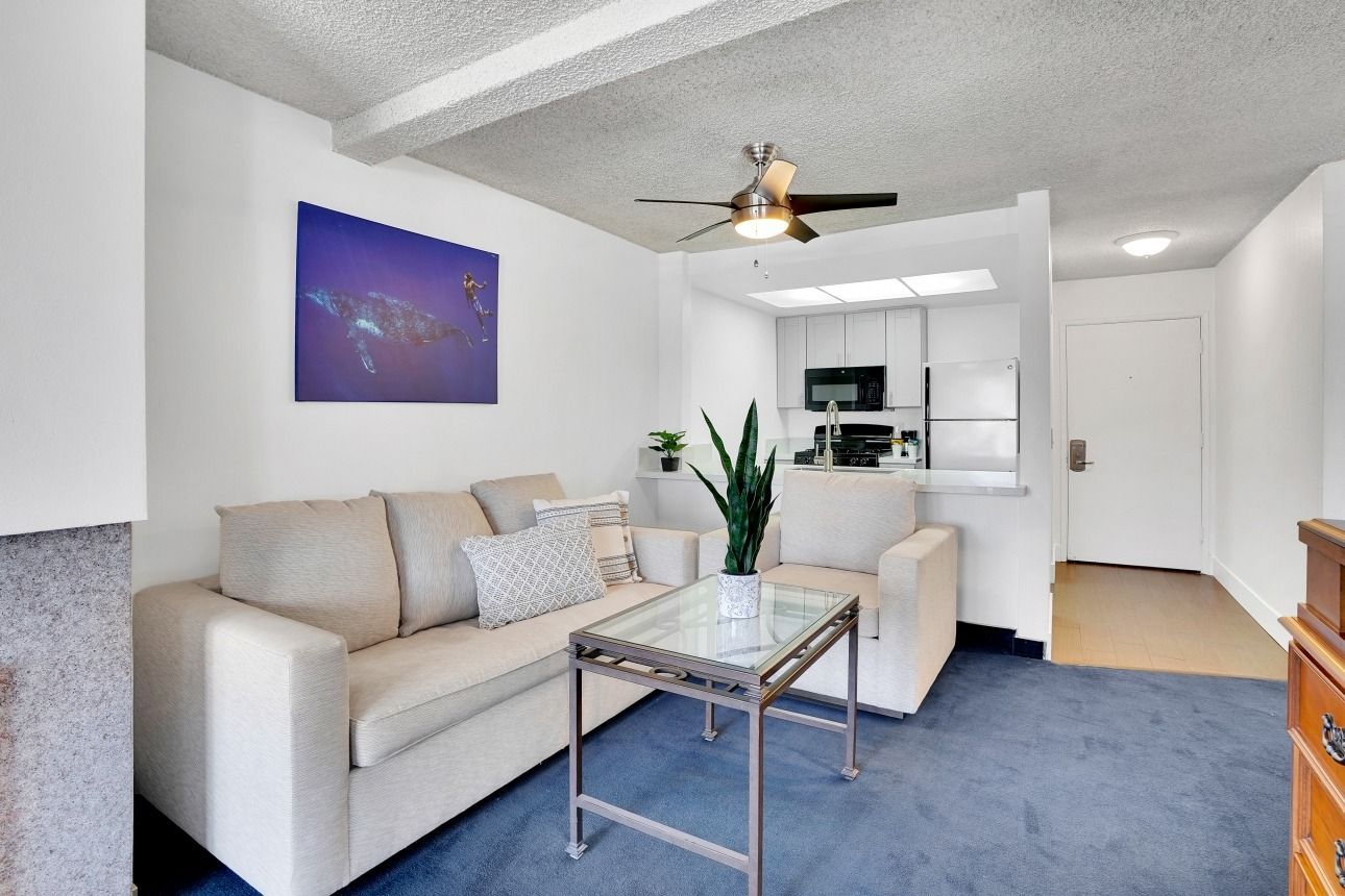 A living room with a beige sofa, glass coffee table, and a houseplant, opening to a white kitchen and a front door.