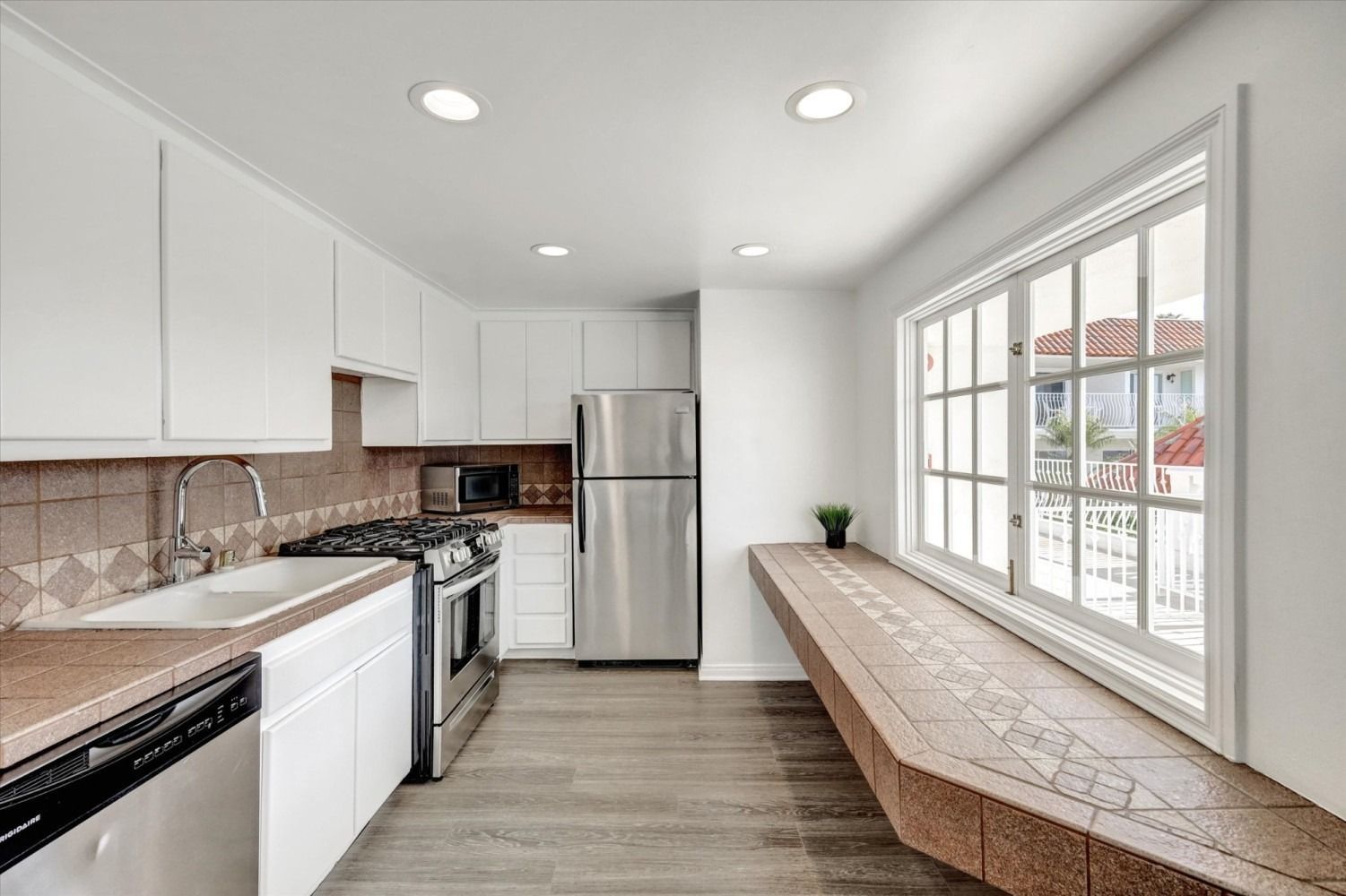 A modern white kitchen featuring stainless steel appliances, wood-look flooring, and a long tile-topped window bench.