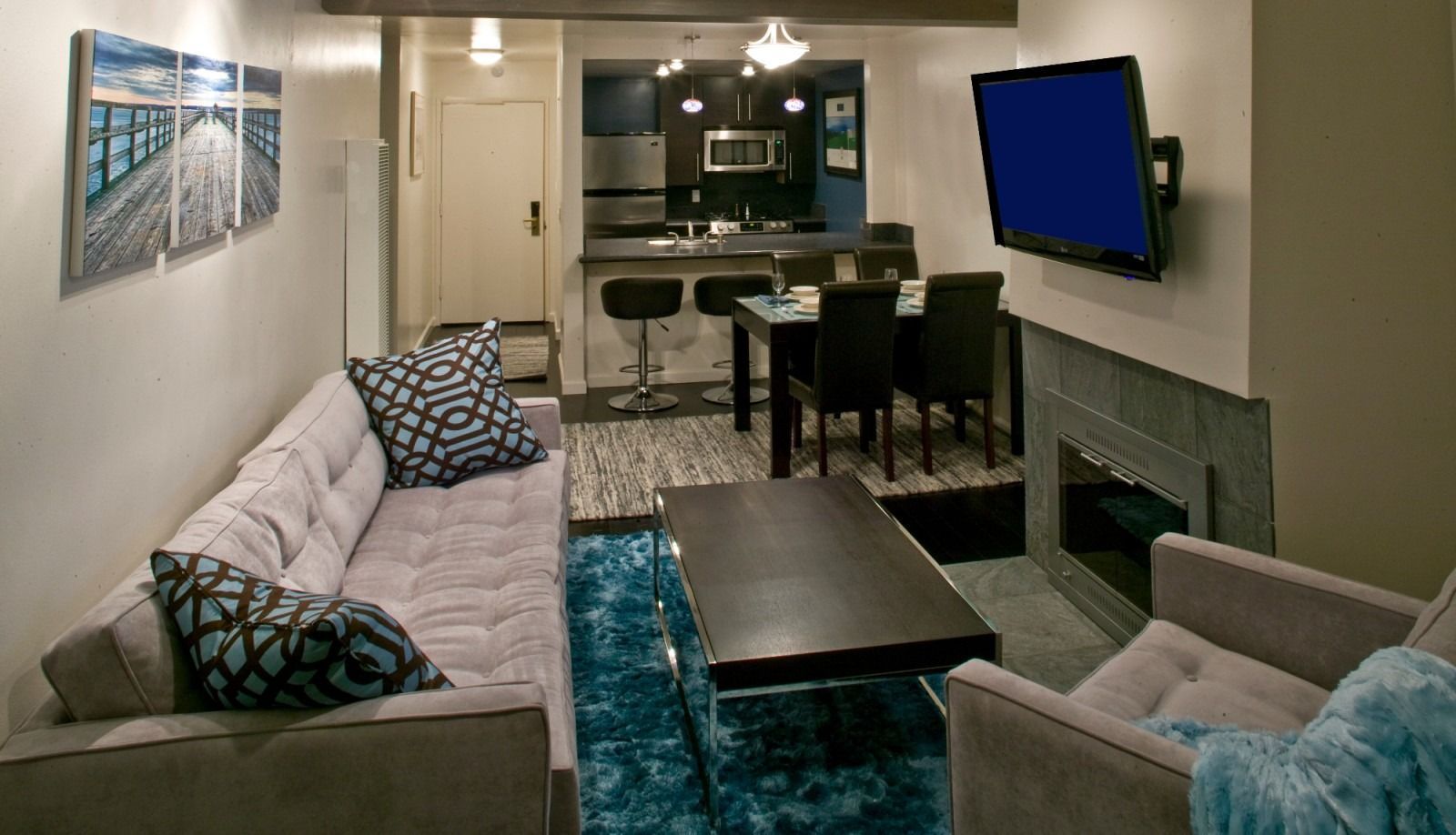 A modern living room with a grey sofa, black coffee table, blue shaggy rug, television, and an open-concept kitchen area.