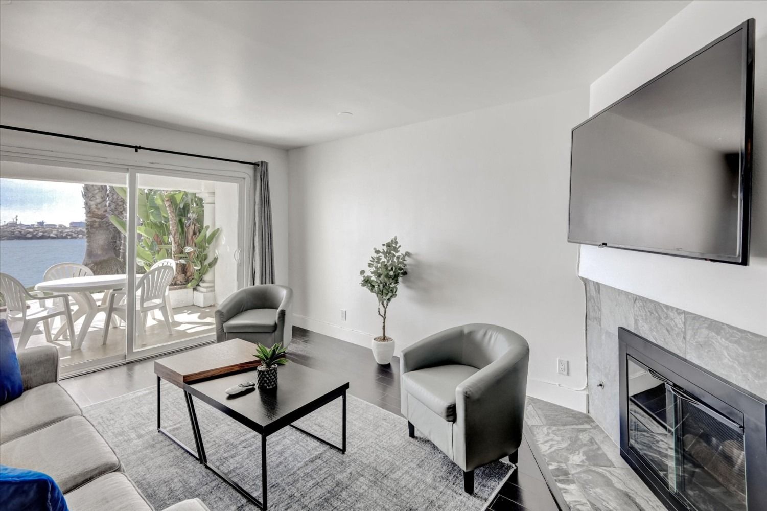 A living room with a gray sofa, two armchairs, a coffee table, a fireplace with a mounted TV, and a view of the water.
