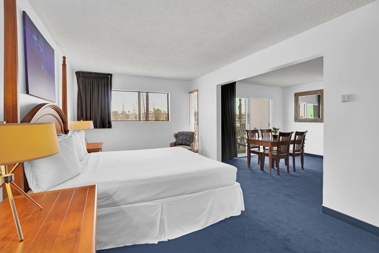 A hotel suite featuring a king bed in the foreground, wooden bedside furniture, and an adjacent dining area with blue carpet.
