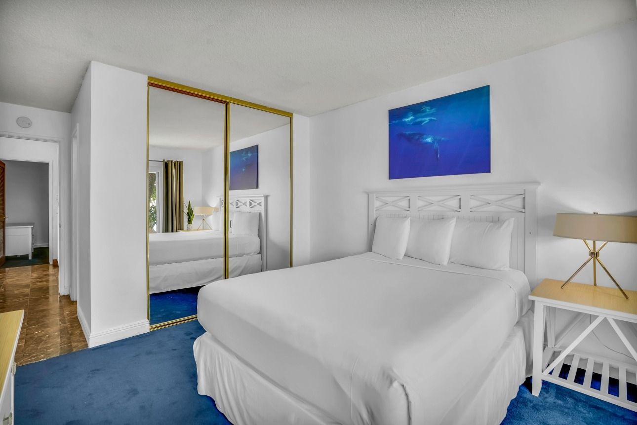 A bedroom with a bed, white bedding, a blue rug, a large mirror closet, and a small side table with a lamp.