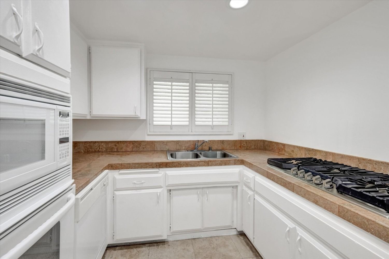 A bright kitchen with white cabinets, tan tiled countertops, a double-basin sink, and a black built-in stovetop.