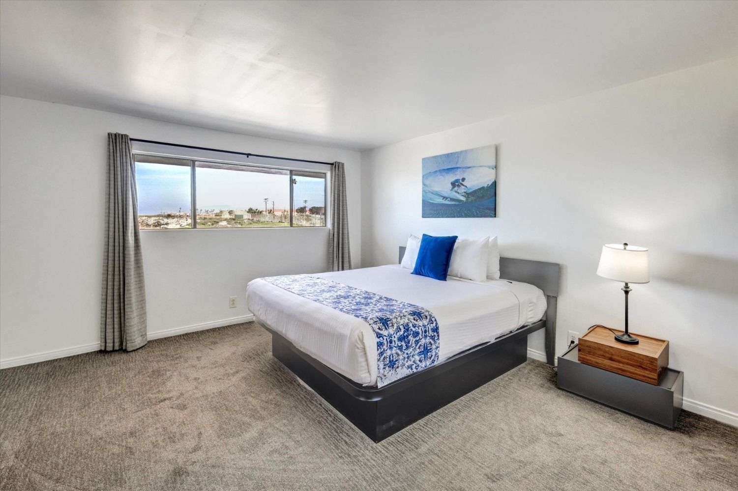 A hotel bedroom featuring a queen bed with a blue patterned runner, white bedding, a desk lamp, and a window with a view.