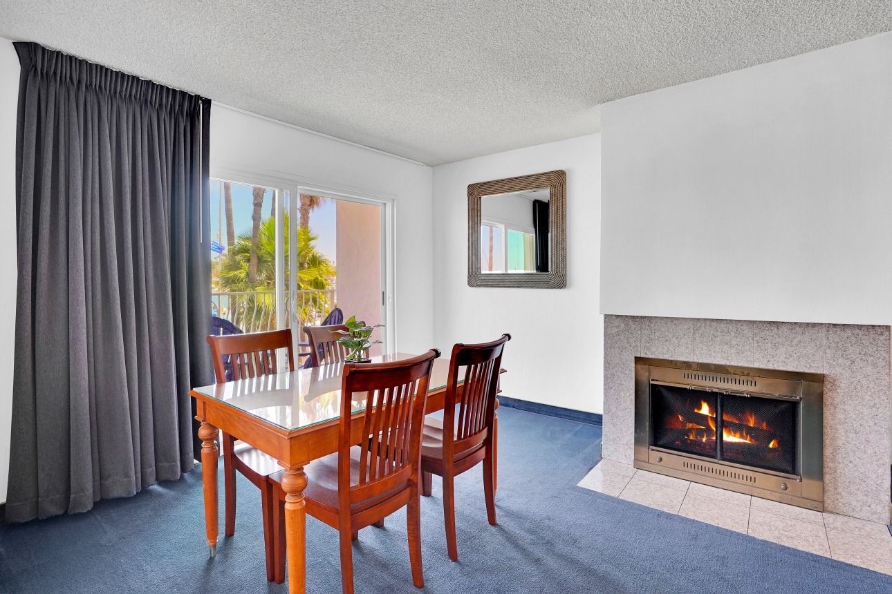 A dining table with four wooden chairs stands in a room with blue carpet, gray curtains, a mirror, and a fireplace.