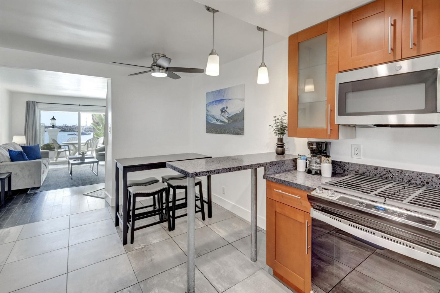 A bright kitchen with wood cabinets, stone countertops, a dining bar with three stools, and a view into a living room.