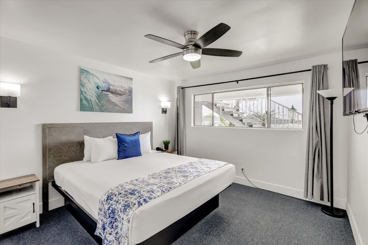 A bedroom with a king bed featuring white bedding, a blue accent pillow, and a wave wall art piece above the headboard.