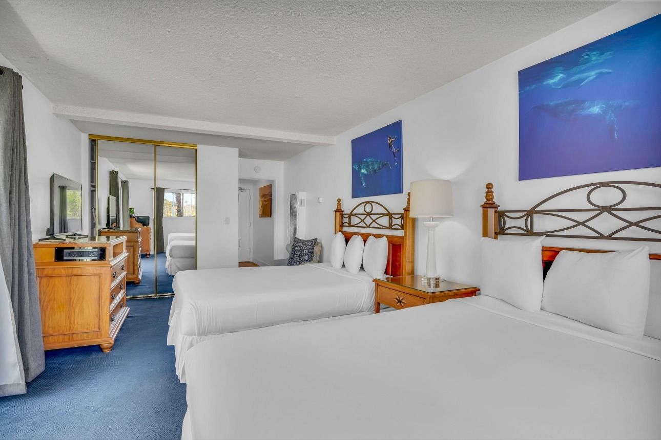 A hotel room with two white beds, blue carpet, wooden dressers, and large ocean-themed art on white walls.