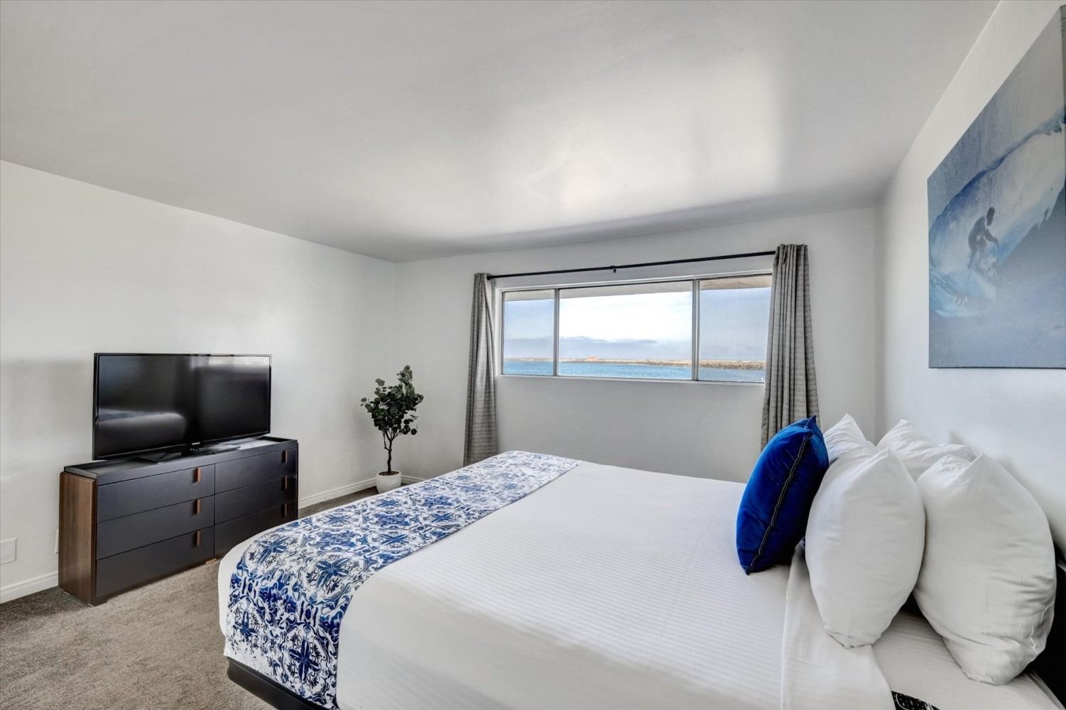 A bright bedroom featuring a queen bed with white linens, a blue runner, and pillows, facing a window with a water view.