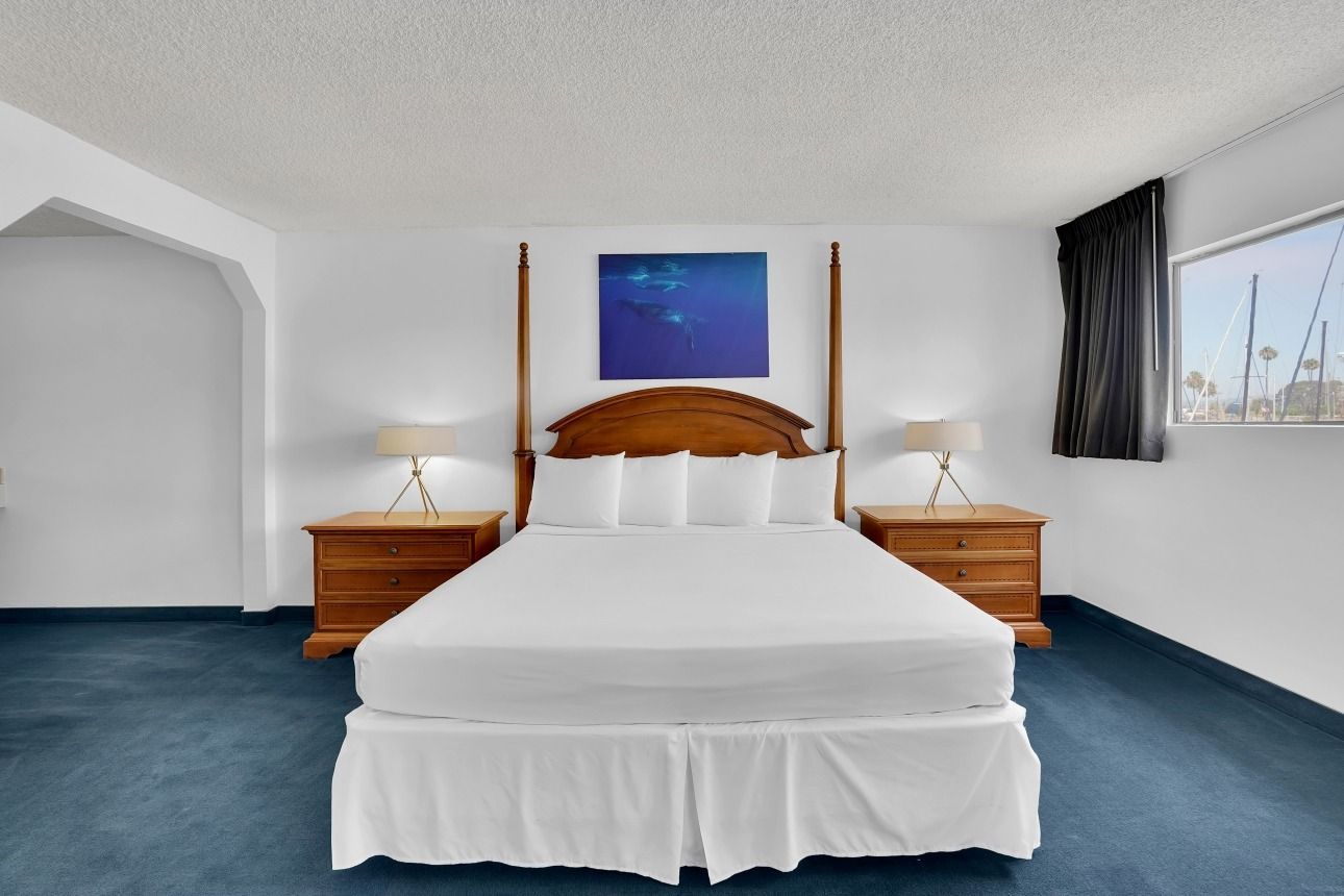 A bedroom with a queen-sized bed, white linens, wooden headboard, two nightstands, blue carpet, and a blue wall painting.