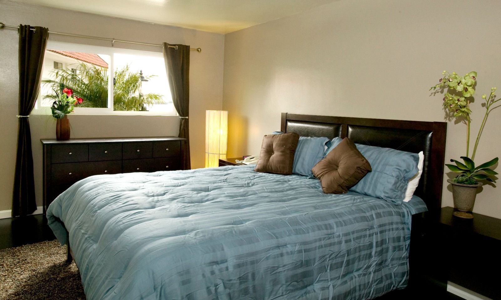 A bedroom with a bed featuring a blue comforter and brown pillows, dark furniture, a window, and a potted plant.