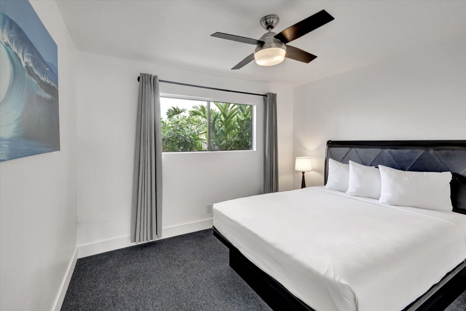 A hotel bedroom with a white-made king bed, grey carpet, a ceiling fan, a window with grey curtains, and ocean wall art.