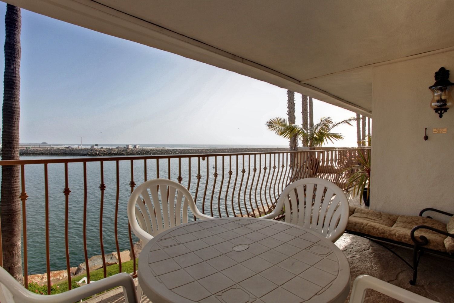 A white patio table and two matching chairs on a balcony overlooking a calm ocean with a stone breakwater in the distance.