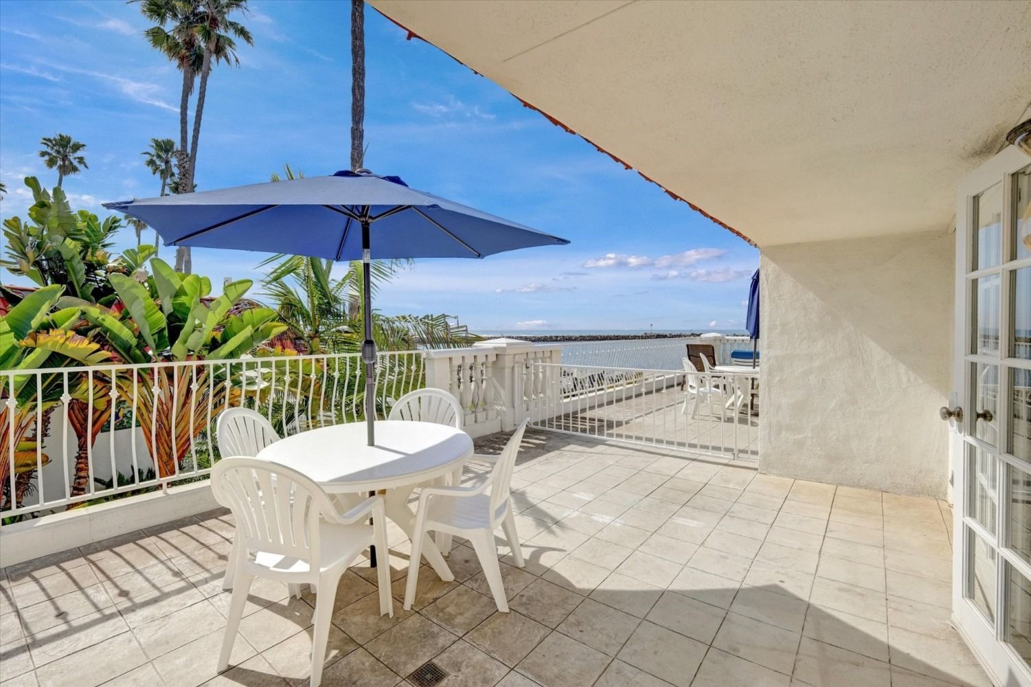 A white patio set sits under a blue umbrella on a tiled balcony overlooking the ocean under a bright, clear sky.