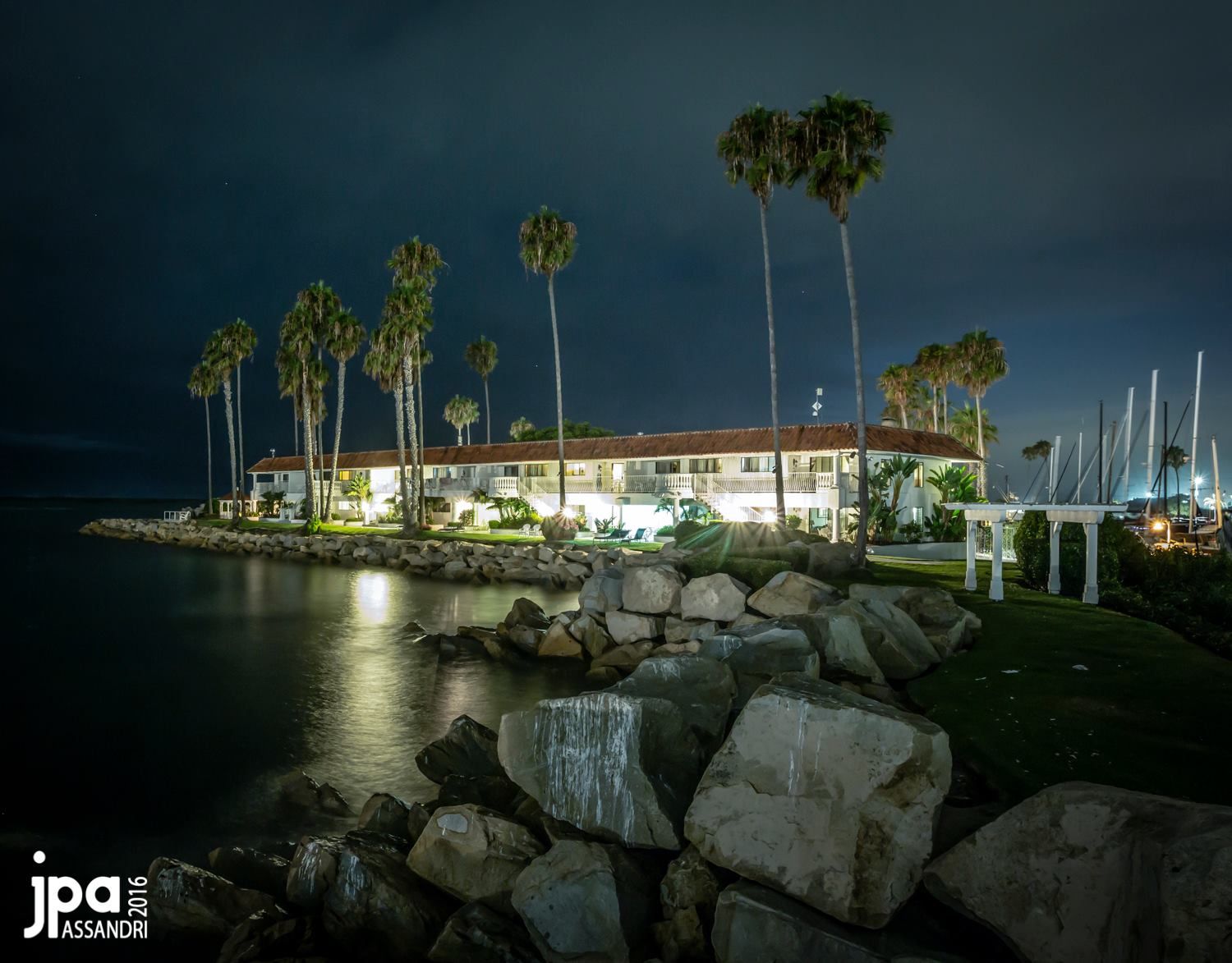 Night view of a coastal building with palm trees, illuminated by lights, against a dark blue sky and calm water.