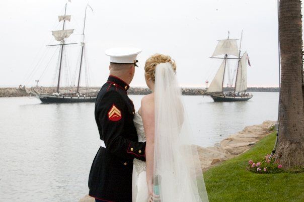 Marine in uniform and bride in veil looking out at two sailboats on water.