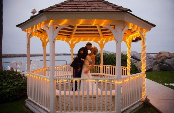 Bride and groom kissing under a gazebo lit with string lights by the water.