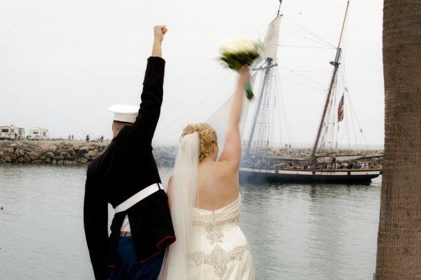 A Marine in uniform and a bride raise arms with fists and bouquet by water, ship in background.